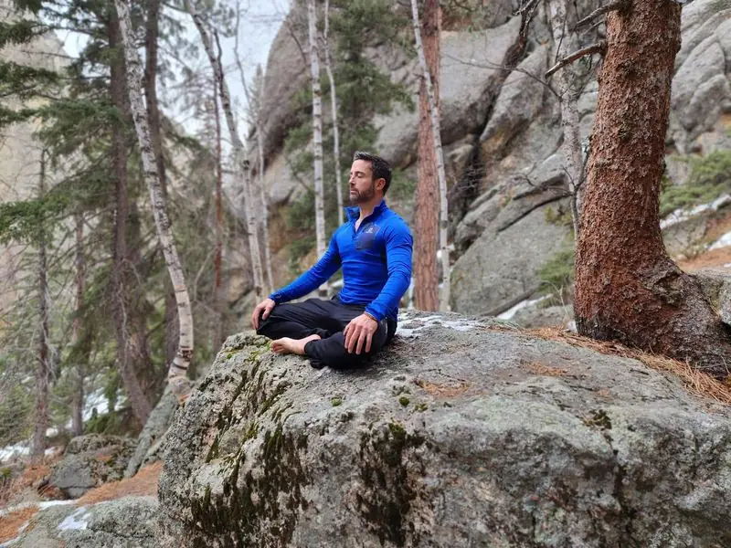 Travis meditating cross-legged on boulder in rocky pine forest