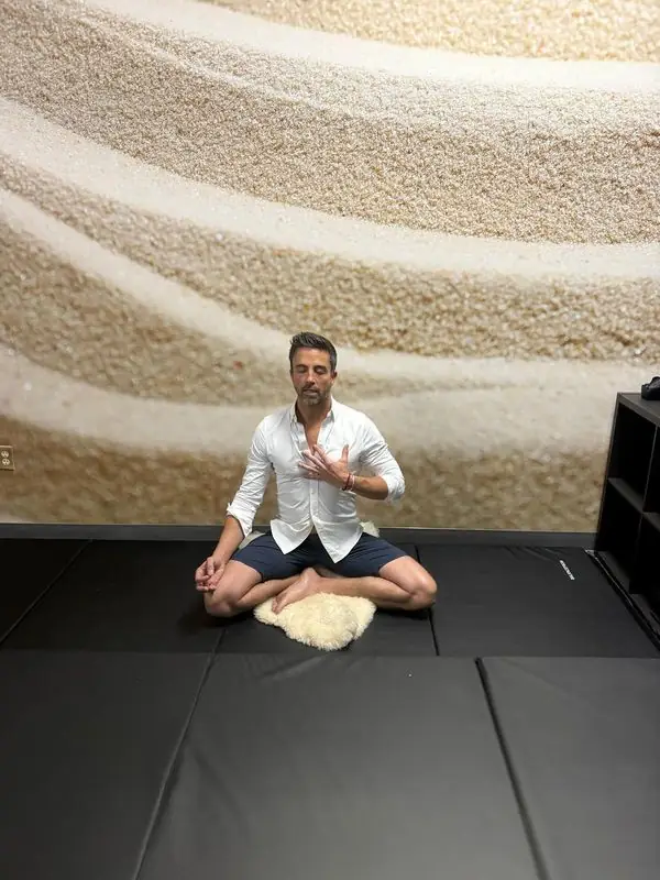 Travis meditating cross-legged with hand on chest, studio mats