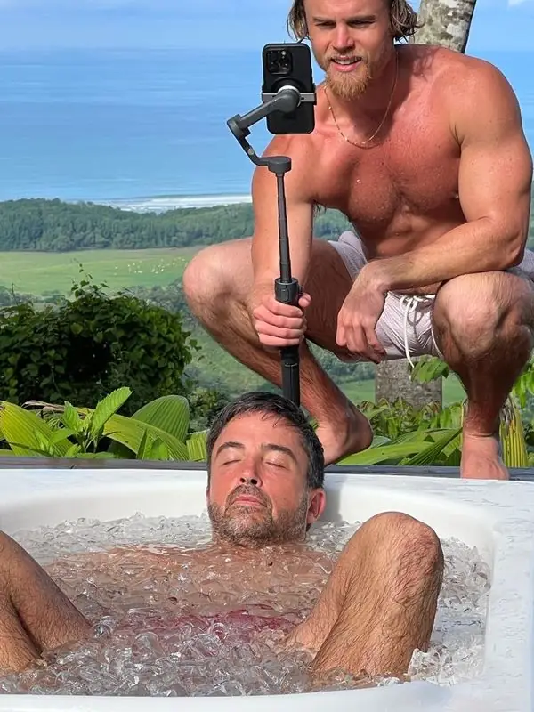 Travis in ice bath eyes closed, friend filming, ocean backdrop