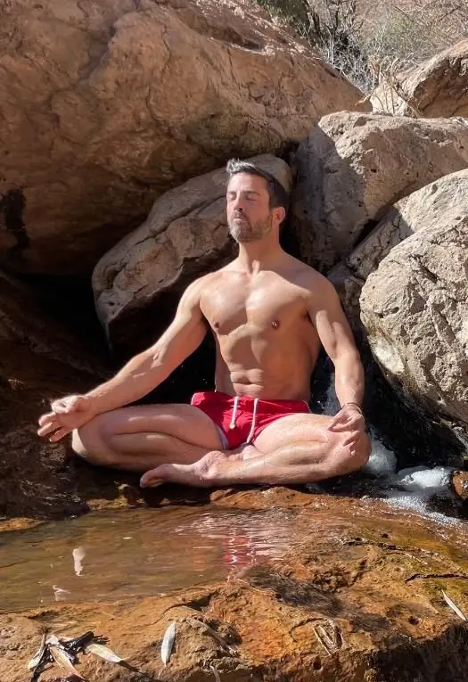 Shirtless lotus-pose meditation on rocks beside small waterfall