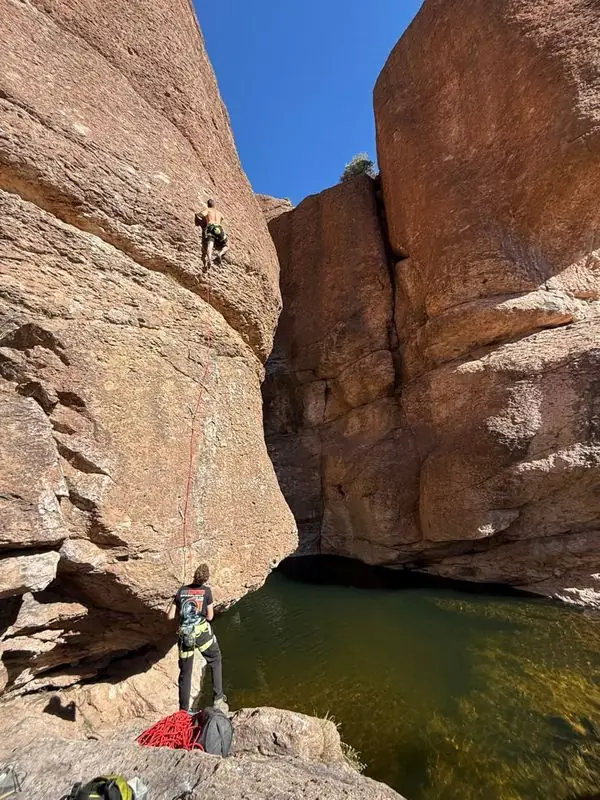 Shirtless climber leading route above canyon swimming hole