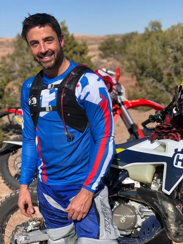 Smiling Travis in blue moto gear beside dirt bikes