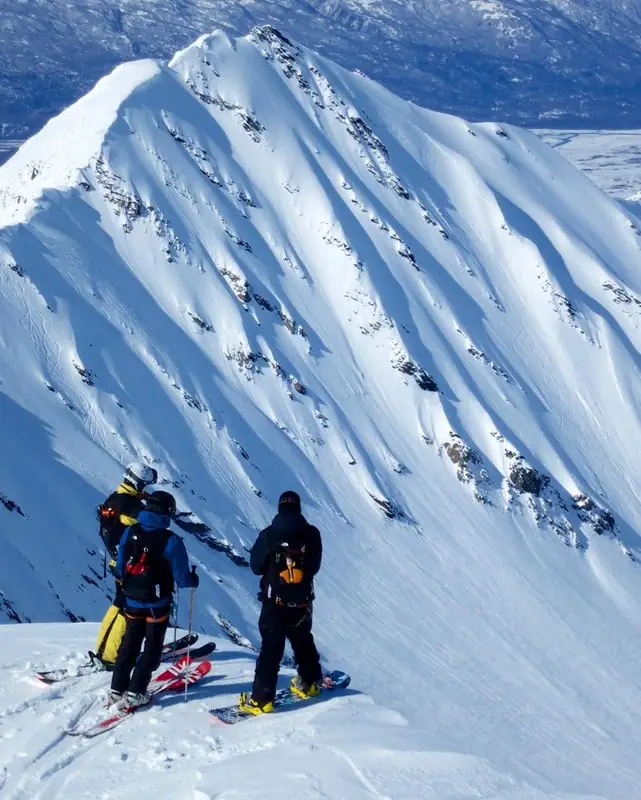 Skiers and snowboarder atop corniced Alaska-style peak